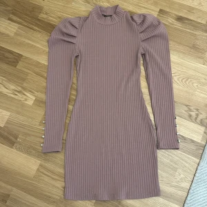 Ribbad lila kortklänning med puffärm - Superfin ribbad lila klänning med puffärmar och höga ärmslut med dekorativa knappar. Klänningen har en tight passform, hög hals och är tillverkad i ett stretchigt material som sitter snyggt på kroppen. Perfekt för dig som gillar trendiga detaljer och stilren look.