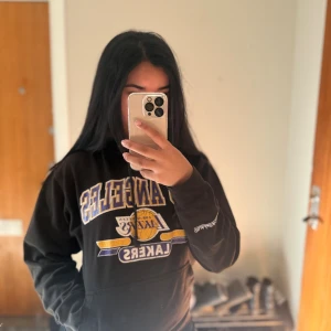 Svart Lakers hoodie från Mitchell & Ness - Svart hoodie med Los Angeles Lakers-tryck i gult, lila och vitt. Stor känguruficka framtill, snörning i huvan och broderad Mitchell & Ness-logga på ärmen. 