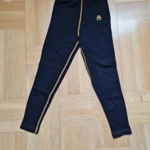 Svarta underställsbyxor från Aclima 100% merinoull 5-6 år - Svarta underställsbyxor från Aclima med kontrasterande gula sömmar och logga på höften. Tight passform och tillverkade i 100%  mjuk merinoull, perfekta för kalla dagar och utomhusaktiviteter. Finns två små hål nära logotypen (se bild). Passar för 5-6 år.