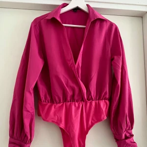 Rosa omlott bodysuit från Lola May - Snygg rosa bodysuit från Lola May med omlott framtill och klassisk krage. Långa ärmar med breda manschetter och klädda knappar. Perfekt att styla med jeans eller kjol för en trendig look.