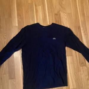 Mörkblå långärmad tröja från Lacoste - Säljer en stilren mörkblå långärmad t shirt från Lacoste med klassisk krokodillogga på bröstet. Tröjan har en rund halsringning och är i regular fit. Nypris 850kr