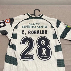 Sporting CP C. Ronaldo #28 fotbollströja - Säljer en klassisk Sporting CP fotbollströja med C. Ronaldo och nummer 28 på ryggen. Tröjan är vit med gröna breda ränder, har korta ärmar och flera snygga tryck, bland annat klubbmärke och sponsorloggor. Materialet är lätt och ventilerande polyester.
