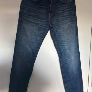 Blå jeans från Jack & Jones, stl 31/34 - Snygga blå jeans från Jack & Jones med klassisk femficksmodell och diskreta slitningar. Jeansen har normal passform och raka ben, perfekta för en avslappnad stil. Tillverkade i slitstarkt denimtyg med kontrastsömmar.