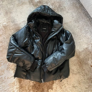 Svart pufferjacka från Pull&Bear - Säljer en svart pufferjacka från Pull&Bear med luva och dragkedja. Jackan har en glansig yta i syntetmaterial som liknar skinn. Perfekt för kalla dagar och riktigt snygg till streetwear. Super varm och vattentät !! Perfekt för hösten men även vintern. 