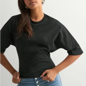 Svart croppad topp med knyt i ryggen - Svart croppad topp i bomull med korta ärmar och rund halsringning. Baksidan har en öppen rygg med breda band som knyts i en snygg rosett. Perfekt för dig som vill ha en cool och trendig look med en twist.