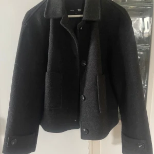 Svart ulljacka från 157 - Stilren svart ulljacka från 157 med klassisk krage, stora knappar framtill och två rejäla fickor. Jackan har en boxig passform och långa ärmar med knappdetalj vid ärmslut. Perfekt för dig som gillar minimalistisk och clean stil.