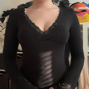 Svart långärmad topp med spets - Svart långärmad topp från Nelly med djup v-ringning och spetskant runt halsen. Figurnära passform och mjukt bomullsmaterial som känns skönt mot huden. Perfekt för dig som gillar en enkel men ändå snygg look.