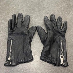 Svarta skinnhandskar från Björn Borg - Svarta skinnhandskar från Björn Borg med snygga silverfärgade dragkedjor vid handleden. Handskarna har ett mjukt stickat foder på insidan för extra värme och komfort. Klassisk design med diskret logga på ovansidan och elastisk mudd för bra passform.