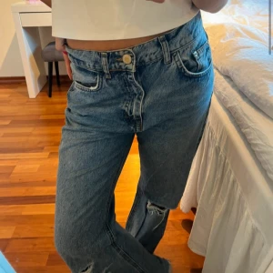 Blå jeans från Pull&Bear - Säljer ett par blå jeans från Pull&Bear med raka ben och snygga slitningar på knä och bakficka. Jeansen har hög midja, klassisk femficksdesign och dragkedja. Perfekta för en avslappnad och trendig look.