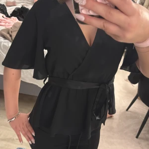 Svart omlottblus med knytband - Säljer en svart omlottblus med v-ringning och korta, fladdriga ärmar. Blusen har ett snyggt knytband i midjan som ger en markerad siluett. Materialet är lätt och luftigt, står inte vart den är ifrån men jag tror SHEIN💕