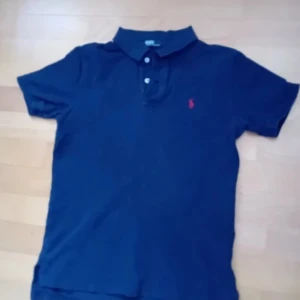 Marinblå pikétröja från Ralph Lauren - Klassisk marinblå pikétröja från Polo Ralph Lauren med korta ärmar och krage. Tröjan har knäppning med tre knappar och den ikoniska rosa broderade loggan på bröstet. Tillverkad i mjuk bomull som är skön att bära hela dagen. Priset är ej hugget i sten 