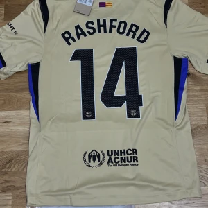  FC Barcelona borta 25/26 # Rashford 14 Player Edition  - Säljer en FC Barcelona fotbollströja med Rashford och nummer 14 på ryggen. Tröjan har korta ärmar, svarta och blå detaljer, samt tryck från Spotify och UNHCR. Materialet är lätt och andas, perfekt för match eller träning. Den är Player Edition en av de högsta kvalitet TVEKA INTE ATT SKICKA EN BUD