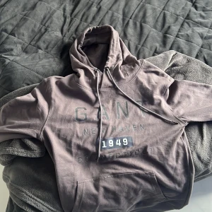 Grå hoodie från GANT med tryck - Säljer en snygg grå hoodie från GANT är från till Tröjan har huva med snörning och en stor magficka. Perfekt för dig som gillar en avslappnad och stilren look.
