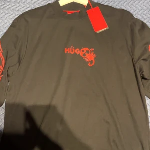 Svart långärmad t-shirt från HUGO - Svart långärmad t-shirt från HUGO med röd tryckt logga och skorpion på bröstet. Ärmarna har unika röda grafiska illustrationer med ansikten, öga och detaljer. T-shirten har rund hals och är gjord i mjuk bomull. Perfekt för dig som gillar statement-plagg och coola prints.Stor i storlek
