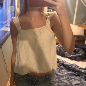 Beige linnetopp med volangdetaljer - Supersöt beige linnetopp med volangdetaljer på axelbanden och spetskant upptill. Toppen är croppad och har en luftig passform, perfekt för varma dagar. Tillverkad i ett lätt och naturligt linnematerial som ger en avslappnad vibe.