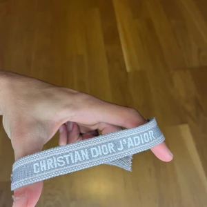Christian Dior J'Adior grå armband - Snyggt grått armband från Christian Dior med texten 'J'Adior' broderad i vitt. Armbandet är gjort i ett mjukt tygmaterial och har fransar vid ändarna för en cool touch. Perfekt accessoar för att lyfta din outfit med lite lyxig Dior-vibe. Pris kan diskuteras fraktar samma dag!