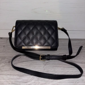 Svar väska - • Svart handväska med gulddetaljer  • Justerbart band • Bandet går att ta av  • Litet fack med dragkedja i väskan