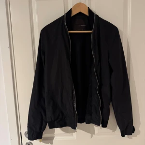 Svart bomberjacka från Zara Man - Stilren svart bomberjacka från Zara Man med klassisk passform. Jackan har dragkedja framtill, ribbade muddar vid ärmslut och midja samt en enkel, clean look utan synliga detaljer. Perfekt för dig som gillar minimalistisk stil.