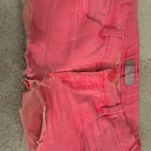Rosa jeansshorts från Jeane Blush - Säljer ett par knallrosa jeansshorts från Jeane Blush. De har råa, fransiga kanter och klassisk femficksdesign. Shortsen är tighta och har knapp- och dragkedjestängning. Perfekta för sommardagar när du vill sticka ut lite extra.