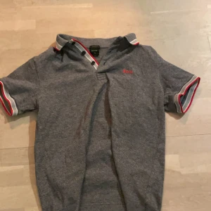 Grå pikétröja från BOSS med röda detaljer - Snygg grå pikétröja från BOSS med klassisk krage, knappar och röda samt vita detaljer på krage och ärmslut. Broderad BOSS-logga i rött på bröstet. Perfekt för en stilren och sportig look.