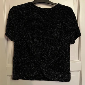 Glittrig svart topp - Säljer en svart glittrig topp från BikBok i storlek M. Toppen har korta ärmar, omlottdetalj framtill