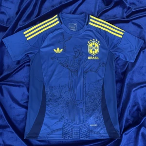 Brasilien fotbollströja  - Snygg blå Brasilien fotbollströja från Adidas med gula detaljer och tre ränder på axlarna. Tryck av Kristusstatyn på framsidan och Brasilien-emblem på bröstet. Tillverkad i lätt och ventilerande material, perfekt för match eller träning.