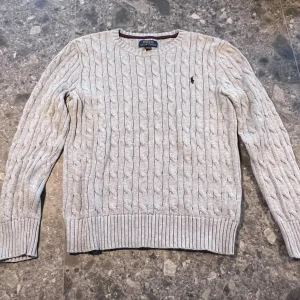 Ljusgrå kabelstickad tröja Polo Ralph Lauren - Kabelstickad tröja från Polo Ralph Lauren i ljusgrått. Klassisk rund halsringning och ribbade muddar vid ärmslut och nederkant. Storlek M (10-12 år).