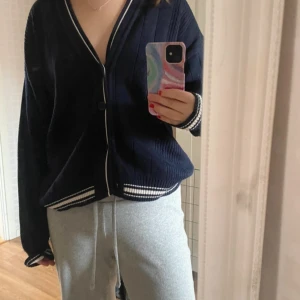 Mörkblå stickad kofta med vita detaljer - Mysig mörkblå stickad kofta med vita ränder längs kanterna och vid ärmsluten. Koftan har v-ringning, knappar framtill och en loose passform. Perfekt att slänga på sig över en t-shirt eller linne för en chill look. Stor i storleken 