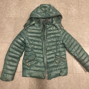 Grön pufferjacka från FJJ XL - Mörkgrön pufferjacka från FJJ i storlek XL. Jackan har huva, två snedställda fickor med guldfärgade dragkedjor och quiltad design. Tillverkad i syntetmaterial och stängs med dragkedja framtill. Perfekt för kalla dagar.