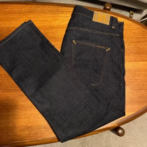 Nudie Jeans Grim Tim Dry True Navy 36/30 - Säljer ett par Nudie Jeans Grim Tim i färgen Dry True Navy. Klassiska jeans med raka ben, snygga kontrastsömmar och femficksdesign. Materialet är 99% bomull och 1% elastan för lite stretch. Perfekta för dig som gillar stilrena och mörkblå jeans.