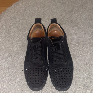Loubs svarta med nitar - Svarta Loubs i mocka med ikonisk röd sula och svarta nitar på tån. Skorna har snörning, rund tå och platt sula. Insidan är i brunt skinn med guldfärgad logga. Perfekt för dig som gillar edgy och lyxig stil. SKRIV VID MINSTA FRÅGA, PRIS KAN DISKUTERAS. 