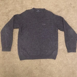 Mörkgrå ulltröja från Henri Lloyd - Mysig mörkgrå stickad tröja från Henri Lloyd med v-ringning och diskret broderad logga på bröstet. Tröjan har långa ärmar och ribbade muddar vid ärmslut och nederkant. Perfekt för kyliga dagar.