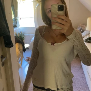 Vit långärmad topp med spetsärmar - Supersöt vit långärmad topp från Lost On Purpose med ribbad struktur, spetsärmar och en liten vinröd rosett vid urringningen. Tryck med logga på ryggen. Perfekt för dig som gillar romantiska detaljer och en feminin vibe.