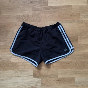 Svarta Adidas träningsshorts - Svarta träningsshorts från Adidas med vita ränder längs sidorna och elastisk midja. Klassisk sportig look med kort modell och lätt material.