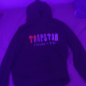 Svart Trapstar hoodie med tryck - Säljer en svart hoodie från Trapstar med stort tryck på bröstet i rött och vitt där det står 'TRAPSTAR IT'S A SECRET'. Klassisk huva och långa ärmar, perfekt för dig som gillar streetwear och vill sticka ut. Pris kan diskuteras