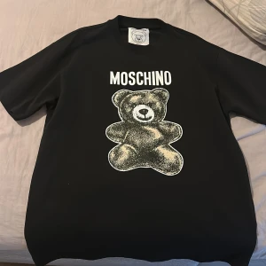 Svart Moschino t-shirt med björntryck - Cool svart t-shirt från Moschino med stort björntryck och logga på bröstet. Tillverkad i 100% bomull för skön känsla. Klassisk passform med korta ärmar och rund hals. Perfekt statement-plagg för dig som gillar designer och streetstyle.