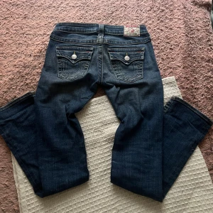 True Religion blå bootcut jeans - Säljer ett par snygga blå jeans från True Religion med bootcut passform. Jeansen har klassiska kontrastsömmar, ikoniska ficklock med knapp och detaljerad brodering på bakfickorna. Materialet är slitstarkt denim och midjan är låg.
