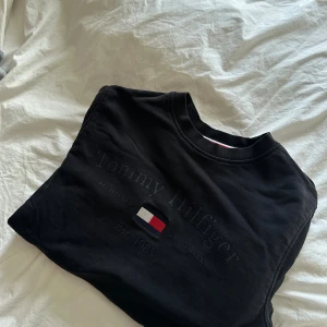 Svart sweatshirt Tommy Hilfiger S - Svart sweatshirt från Tommy Hilfiger med broderad logga och text på bröstet. Klassisk rund halsringning med ribbade muddar och diskreta Hilfiger-detaljer. Tröjan är i mjuk bomull och har lång ärm, perfekt för en clean och stilren look.