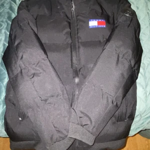 Svart pufferjacka från Tommy Hilfiger - Svart pufferjacka från Tommy Hilfiger med klassisk logga på bröstet. Jackan har dragkedja framtill, ribbade muddar och glansigt svart innerfoder. Perfekt för kalla dagar och har en clean, stilren look.