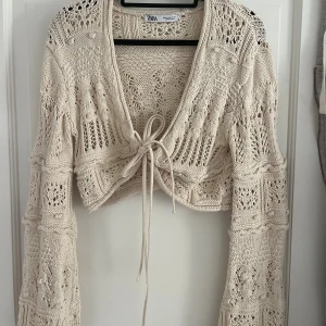 Beige virkad bolerotopp från Zara - Beige bolerotopp från Zara med virkat mönster och knyt framtill. Croppad modell med långärmade ärmar och vida armar.