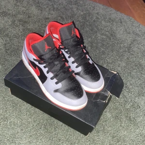 Nike Air Jordan 1 Low svart/röd/grå - Nike Air Jordan 1 Low sneakers med svart och grå ovandel i skinn, röda detaljer och röd insida. Klassisk Jordan-logga på hälen och röd swoosh på sidan. Svarta snören och perforerad tå för extra stil. Snygg och ikonisk siluett som sticker ut.