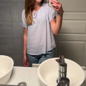 Randig blus med knyt i halsen - Säljer en luftig blus med ljusblå och vita ränder. Blusen har korta ärmar, rund halsringning med knyt och är tillverkad i ett mjukt bomullsliknande material. Perfekt för varmare dagar och enkel att styla med jeans eller kjol.