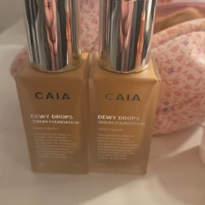 CAIA Dewy Drops Serum Foundation - Två flaskor CAIA Dewy Drops Serum Foundation med silvriga lock. Foundationen har en varm beige ton och innehåller vitamin E och glycerin för återfuktning. Kommer i stilrena fyrkantiga glasflaskor med pump. Perfekt för en fräsch och naturlig look.