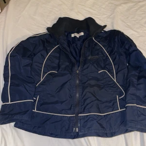 Marwin mörkblå  vindjacka med vita detaljer - Blå vindjacka från Marwin med vita kontrastlinjer och dragkedja framtill. Jackan har hög krage och två fickor på sidorna. Perfekt för kyliga dagar och enkel att matcha med det mesta. Snygg sportig look och bekväm passform.