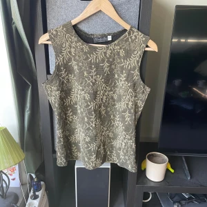 Guldmönstrad ärmlös topp från Number One - Snygg ärmlös topp från Number One by KappAhl i svart med guldigt blommönster. Toppen har rund hals och är tillverkad i ett glittrigt syntetmaterial som ger en lyxig känsla. Perfekt för dig som vill sticka ut med coola detaljer och metallic-vibe.