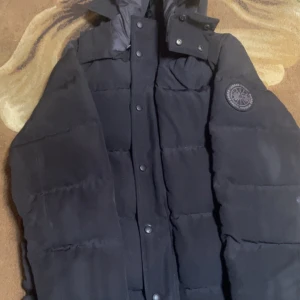 Svart pufferjacka från Canada Goose - Säljer en svart pufferjacka från Canada Goose med hög krage och huva. Jackan har ett stilrent, quiltat mönster och logotyp på bröstet. Perfekt för kalla dagar med sin tjocka och isolerande design. Stängs med både dragkedja och knappar framtill.