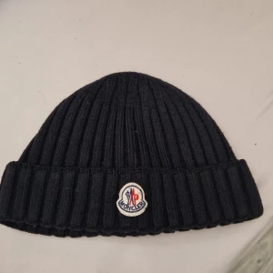 Moncler mössa svart - Svart ribbstickad mössa från Moncler med klassisk uppvikt kant och broderad logotyp framtill. Tillverkad i mjukt material som håller dig varm och snygg under kyliga dagar. Priset går att diskutera!