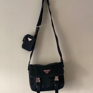 Svart axelväska från Prada med red logga - Snygg svart axelväska från Prada i nylon med klassisk röd triangel-logga framtill. Väskan har justerbar axelrem, silverfärgade spännen och en matchande liten pouch med dragkedja. Perfekt för dig som vill ha en stilren och lyxig accessoar. original price: 21k sek. receipt is available on request! meet up and swish ready