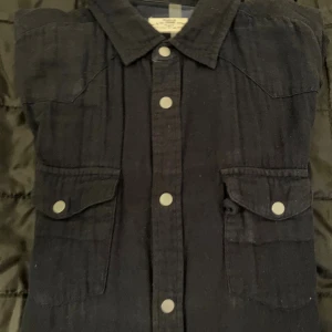 Mörkblå overshirt med knappar - Snygg mörkblå overshirt från RRL Ralph Lauren med klassisk krage, tryckknappar och två bröstfickor med lock. Långärmad modell i mjuk bomull som ger en chill och stilren vibe. Perfekt att bära som lager över t-shirt eller hoodie.