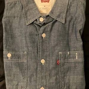 Blå overshirt från Levi's i bomull - Snygg blå overshirt från Levi's i storlek M, tillverkad i mjuk bomull. Skjortan har klassisk krage, vita knappar framtill och två bröstfickor med tydliga sömmar. Perfekt för lager-på-lager och chill stil. Levi's logga syns på ena fickan.